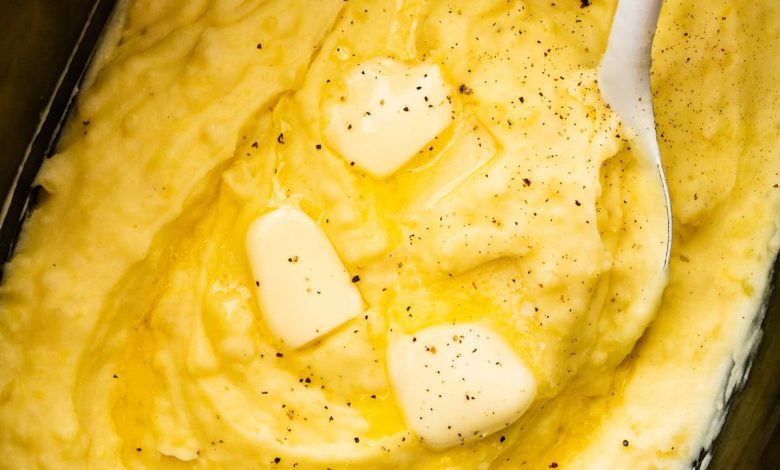 Mashed potatoes in a slow cooker with slices of butter, seasonings, and a spatula.