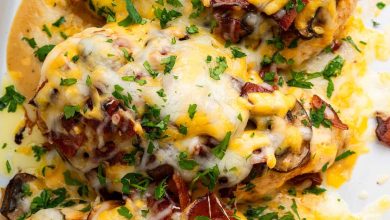 Four chicken breasts in a white baking dish topped with mushrooms, turkey bacon, melted cheese and fresh parsley.