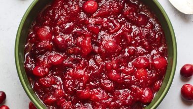 A serving dish containing cranberry sauce. A spoon rests at the top of the bowl and there are whole cranberries scattered around the bowl.