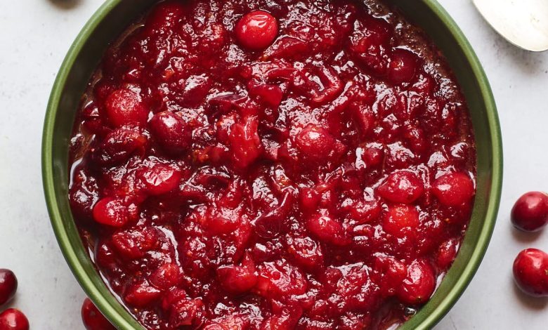 A serving dish containing cranberry sauce. A spoon rests at the top of the bowl and there are whole cranberries scattered around the bowl.