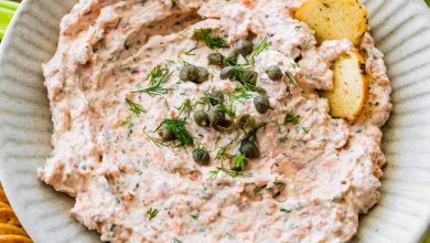 A bowl of smoked salmon dip, garnished with fresh dill and capers, served with crackers and fresh vegetables.