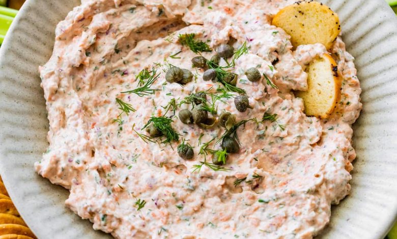 A bowl of smoked salmon dip, garnished with fresh dill and capers, served with crackers and fresh vegetables.