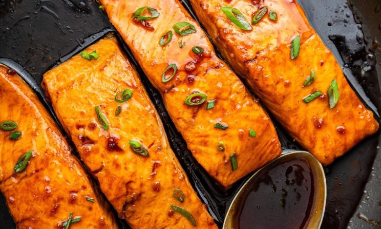 Four honey glazed salmon filets garnished with fresh chives in a skillet with a metal spoon that contains some of the honey based sauce in it.