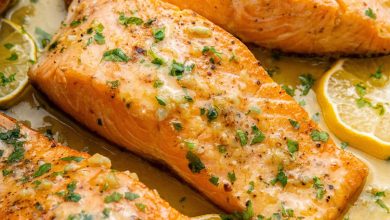 Four salmon filets in a pan with garlic butter and lemon wedges. The filets are garnished with fresh herbs.