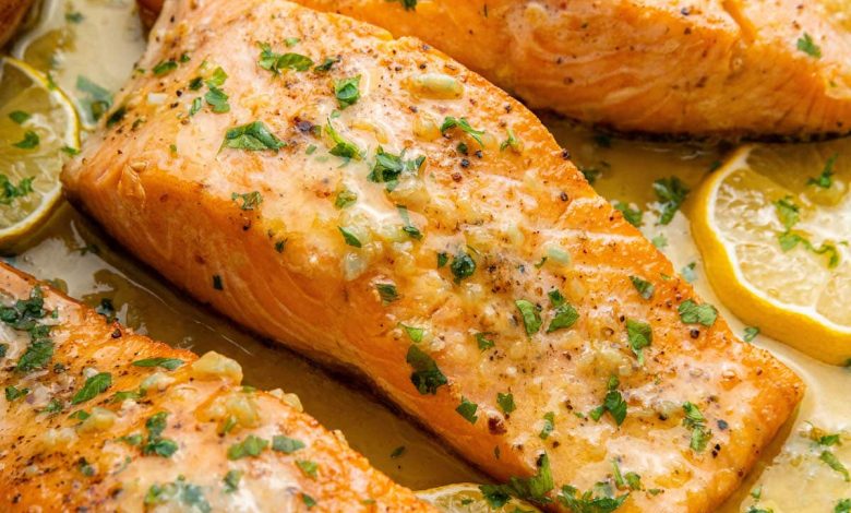 Four salmon filets in a pan with garlic butter and lemon wedges. The filets are garnished with fresh herbs.