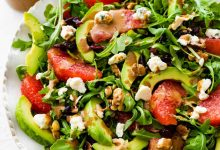 A salad with arugula, avocado, blue cheese, grapefruit, nuts, and cranberries on a white plate.