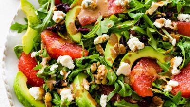 A salad with arugula, avocado, blue cheese, grapefruit, nuts, and cranberries on a white plate.