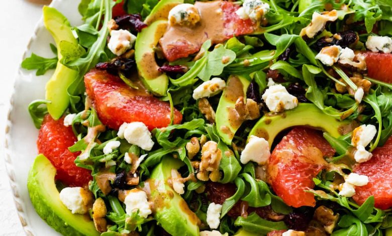 A salad with arugula, avocado, blue cheese, grapefruit, nuts, and cranberries on a white plate.