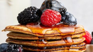 Maple syrup being poured on top of a stack of six greek yogurt pancakes on a plate. They