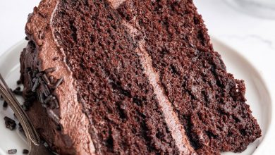 A slice of chocolate cake with chocolate buttercream frosting on a white plate with a metal fork to the side.