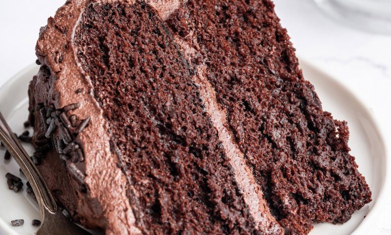 A slice of chocolate cake with chocolate buttercream frosting on a white plate with a metal fork to the side.