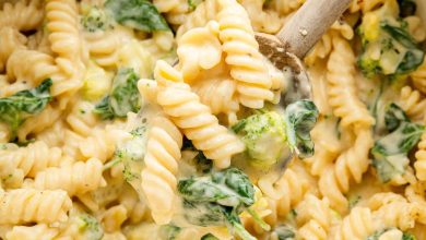 Rotini pasta, broccoli and spinach in a pot coated in a cheese sauce. A wooden spoon is scooping up some of the pasta.