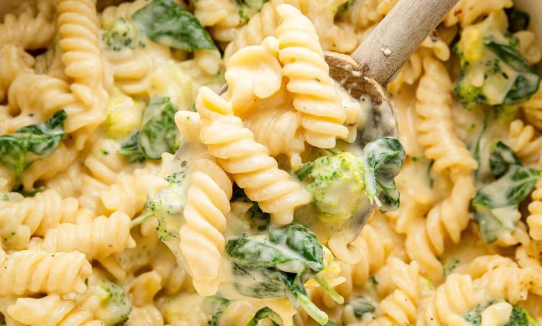 Rotini pasta, broccoli and spinach in a pot coated in a cheese sauce. A wooden spoon is scooping up some of the pasta.