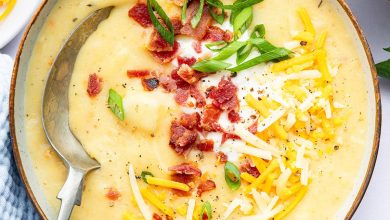 A bowl of potato soup with a silver spoon in it. The soup is topped with bacon bits, green onion, shredded cheese and sour cream.