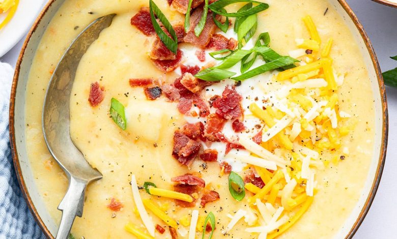 A bowl of potato soup with a silver spoon in it. The soup is topped with bacon bits, green onion, shredded cheese and sour cream.