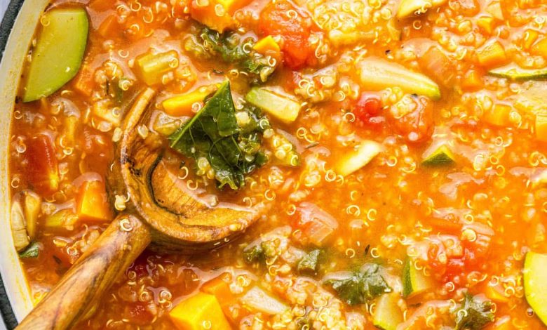 A large pot of vegetable packed quinoa soup with a wooden serving spoon in the soup.