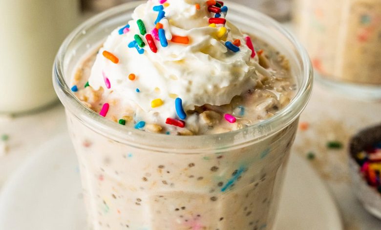 A glass jar filled with birthday cake overnight oats, topped with whipped cream and colorful sprinkles, with a bottle of milk and another jar of oats in the background.