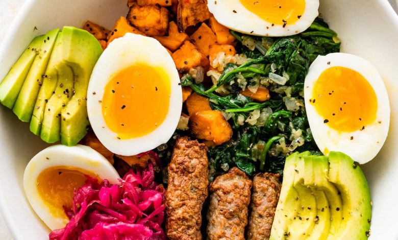 A breakfast bowl with 2 soft boiled eggs, 3 turkey sausage, sliced avocado, sauerkraut, roasted sweet potatoes and sautéed kale all in a white bowl.