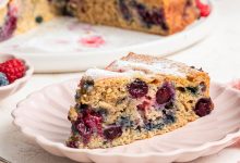 A slice of mixed berry cake on a light pink plate. The entire berry cake is in the background on a white plate.