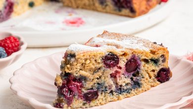 A slice of mixed berry cake on a light pink plate. The entire berry cake is in the background on a white plate.