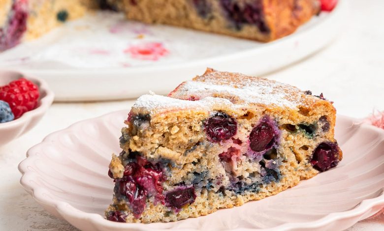 A slice of mixed berry cake on a light pink plate. The entire berry cake is in the background on a white plate.