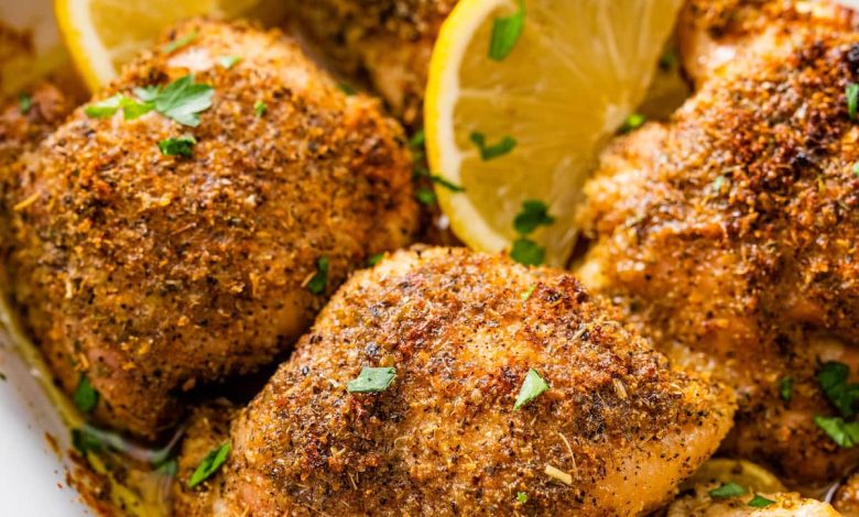 Six baked Greek chicken thighs in a white baking dish. The chicken is topped with fresh parsley and lemon slices.