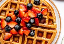 Golden brown protein waffles on a plate, topped with fresh strawberries and blueberries, drizzled with syrup, and served with a side of fruit.