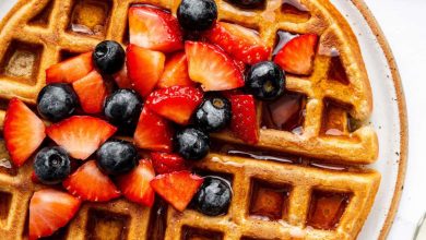 Golden brown protein waffles on a plate, topped with fresh strawberries and blueberries, drizzled with syrup, and served with a side of fruit.