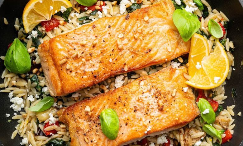 Two salmon filets in a skillet served over a bed of orzo combined with roasted red peppers, chopped dates, feta and swiss chard. It