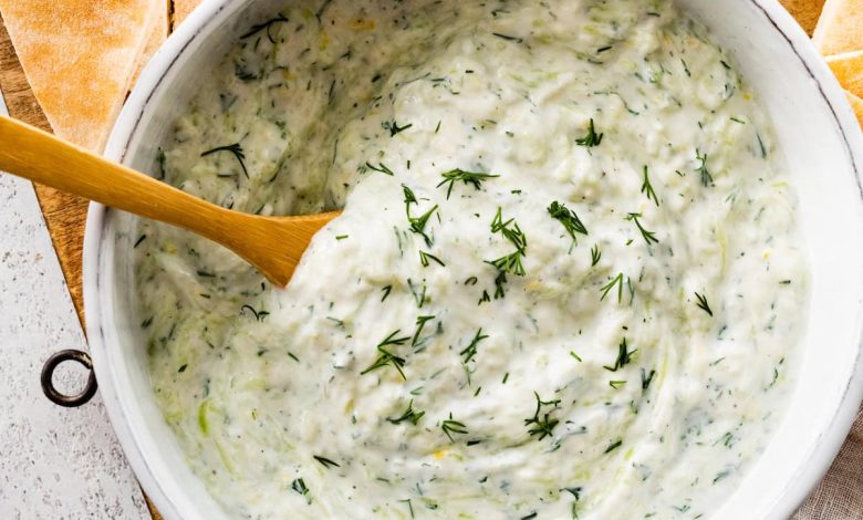 A white bowl of tzatziki topped with fresh dill. There is a wooden spoon in the bowl and the bowl is surrounded by pita bread and lemon slices.