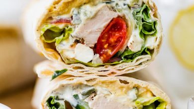 Two halves of a Greek chicken wrap stacked on top of each other on a wooden cutting board. You can see the tomatoes, chicken, lettuce and sauce inside the wrap.