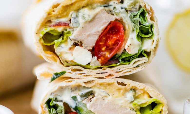 Two halves of a Greek chicken wrap stacked on top of each other on a wooden cutting board. You can see the tomatoes, chicken, lettuce and sauce inside the wrap.