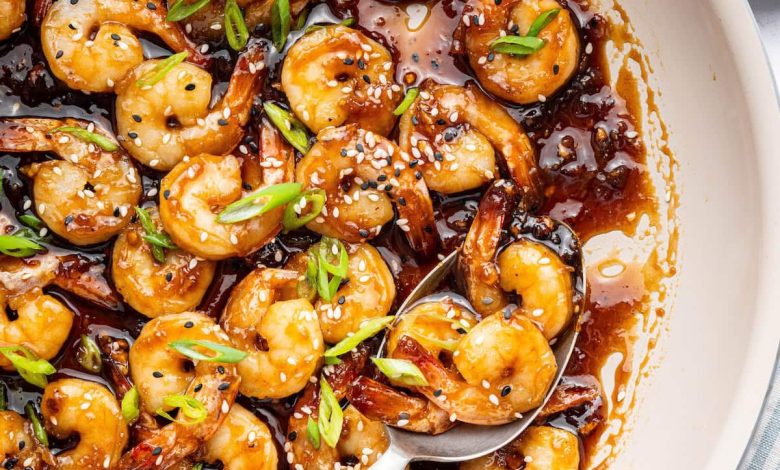 Shrimp coated in a honey garlic sauce in a skillet. There is a silver spoon in the skillet and the shrimp is topped with sesame seeds and green onion slices.