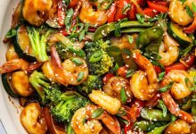 Shrimp stir-fry with shrimp, broccoli, red bell peppers, snap peas, zucchini, and onions coated in sauce. The dish is garnished with sesame seeds and chopped green onions, with a wooden spoon in the pan.