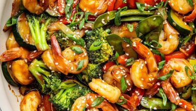 Shrimp stir-fry with shrimp, broccoli, red bell peppers, snap peas, zucchini, and onions coated in sauce. The dish is garnished with sesame seeds and chopped green onions, with a wooden spoon in the pan.