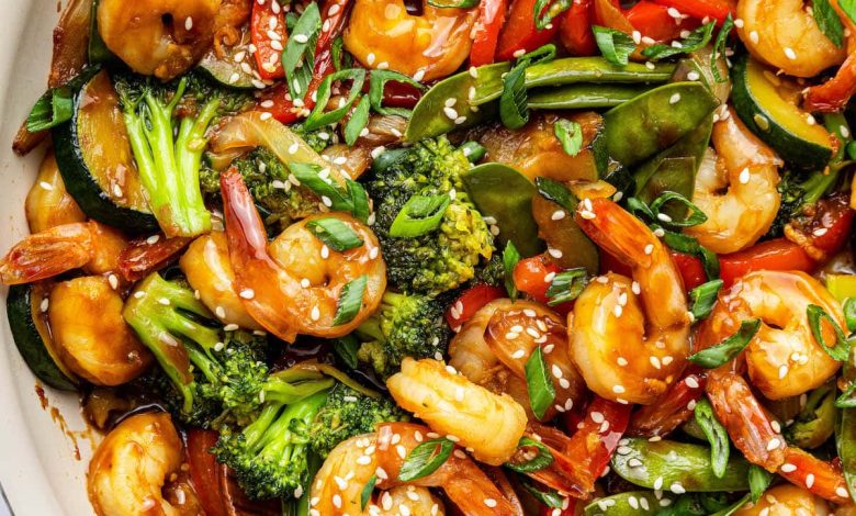 Shrimp stir-fry with shrimp, broccoli, red bell peppers, snap peas, zucchini, and onions coated in sauce. The dish is garnished with sesame seeds and chopped green onions, with a wooden spoon in the pan.