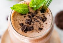 One mint chocolate protein shake in a glass topped with crushed Thin Mint cookies and fresh mint. There is a green straw in the glass.