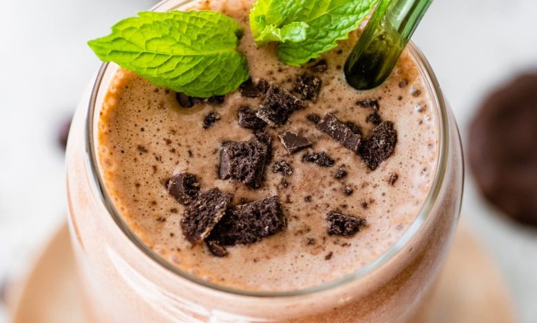 One mint chocolate protein shake in a glass topped with crushed Thin Mint cookies and fresh mint. There is a green straw in the glass.