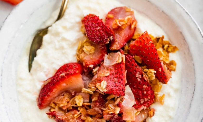 Cottage cheese in a bowl topped with roasted strawberries, rolled oats and coconut. There is a silver spoon in the bowl.