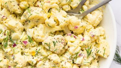 Cauliflower potato salad in a large serving bowl with silver serving utensils. The salad is topped with fresh dill.