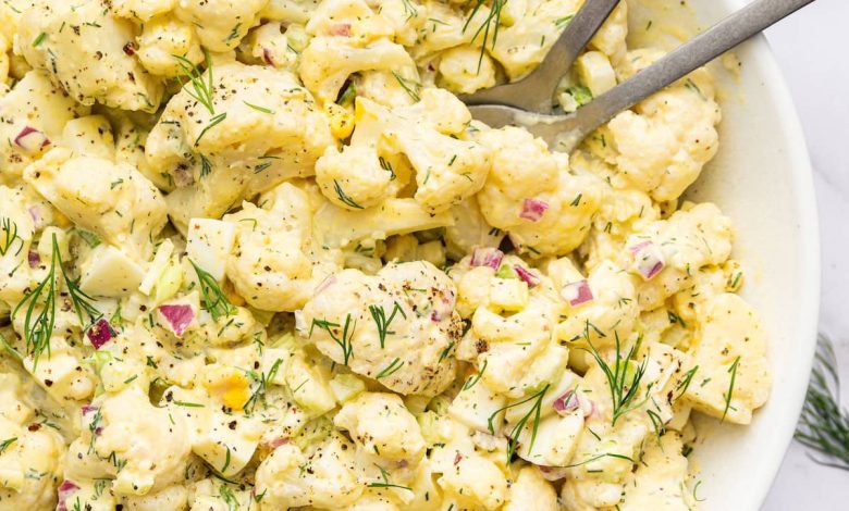 Cauliflower potato salad in a large serving bowl with silver serving utensils. The salad is topped with fresh dill.