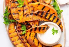 Grilled sweet potatoes on a serving platter topped with fresh parsley and served with a side of garlic aioli. There is one sweet potato dipped in the aioli.