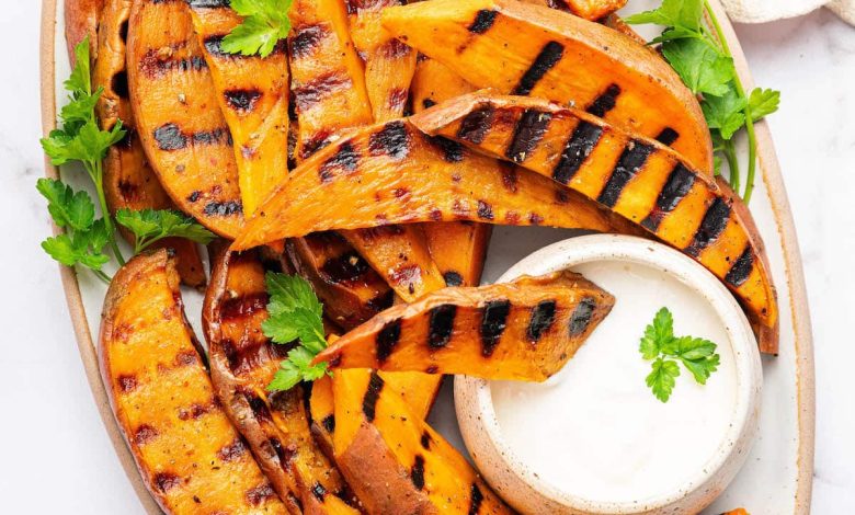 Grilled sweet potatoes on a serving platter topped with fresh parsley and served with a side of garlic aioli. There is one sweet potato dipped in the aioli.