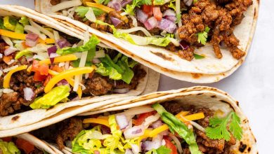 Five ground beef tacos in flour tortillas topped with lettuce, onion, tomato, cilantro and cheese.