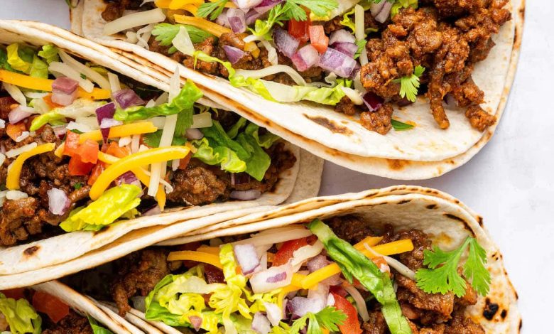 Five ground beef tacos in flour tortillas topped with lettuce, onion, tomato, cilantro and cheese.