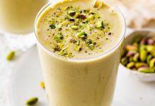 One glass filled with a pistachio smoothie topped with crushed pistachios. There is a clear straw in the glass and another smoothie in the background.