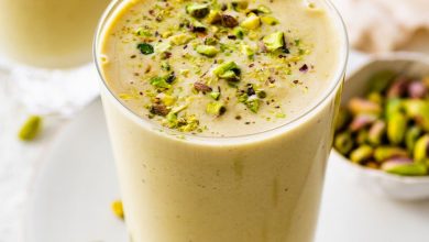 One glass filled with a pistachio smoothie topped with crushed pistachios. There is a clear straw in the glass and another smoothie in the background.