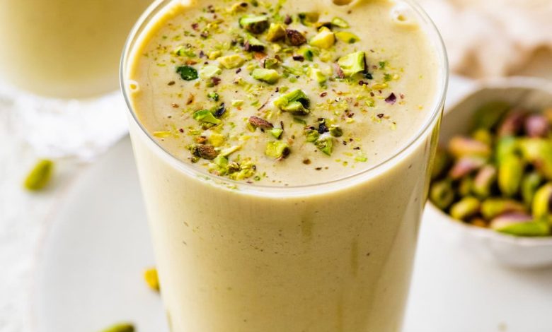 One glass filled with a pistachio smoothie topped with crushed pistachios. There is a clear straw in the glass and another smoothie in the background.