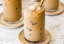 A glass of iced coffee with milk fully mixed in, served on a small wooden plate with a metal straw.