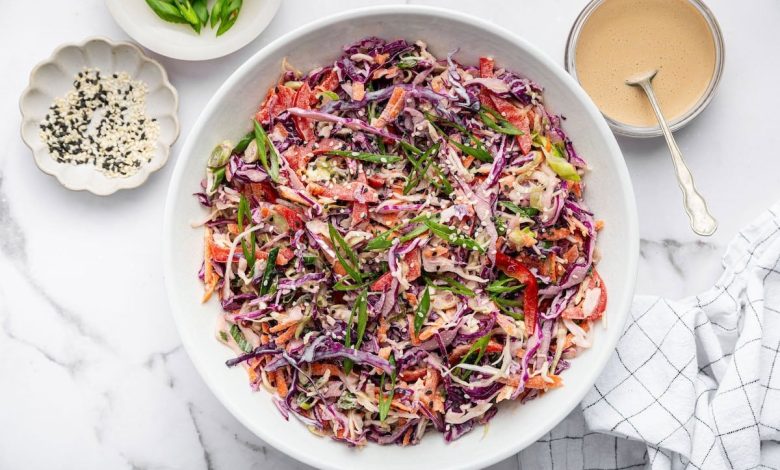 Assembled Asian slaw in a white bowl, garnished with sesame seeds and scallions. Dressing and toppings sit around the bowl on a marble counter with a striped towel.